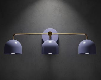 May include: A modern wall-mounted light fixture with three lavender-colored dome shades. The fixture has a gold-colored horizontal bar and a circular lavender base, mounted on a dark gray wall. The light fixture is designed for interior use.