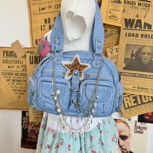 May include: A light blue handbag with multiple pockets, adorned with a leopard print star and silver chain details. The bag features a top handle and a long chain with cross and pearl accents. The background includes vintage newspaper clippings.
