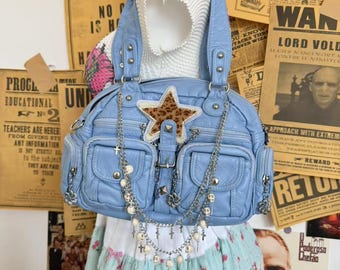 May include: A light blue handbag with multiple pockets, adorned with a leopard print star and silver chain details. The bag features a top handle and a long chain with cross and pearl accents. The background includes vintage newspaper clippings.