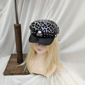 Puede incluir: Una gorra de estilo marinero con un estampado de leopardo en blanco y negro en la parte superior. La gorra incluye una visera negra y una banda negra adornada con tachuelas metálicas plateadas y un emblema central de calavera plateada.