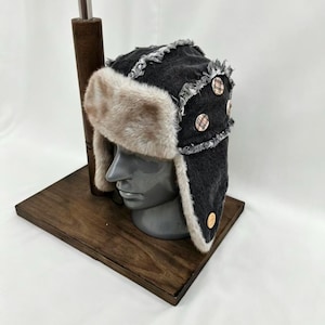 May include: A dark gray denim trapper hat featuring a thick, light brown faux fur lining and trim. The hat includes frayed seam detailing and three circular buttons with a tan and brown plaid pattern attached to the side ear flap section.