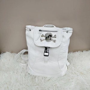 May include: A white backpack featuring a textured surface, a top flap with a silver-toned skull emblem adorned with clear crystals, a zippered pocket on the flap, and a black plastic buckle closure on the front center of the bag.