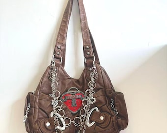 May include: Brown leather shoulder bag with silver chain accents. The bag features a red heart-shaped emblem with the word "TAKEN" and a keyhole design. Additional silver embellishments include the letters "S" and "C".
