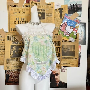 Peut inclure: Un haut de style dos nu à motifs floraux aux couleurs pastel, orné de dentelle et de volants blancs. Le haut est présenté sur un mannequin devant un mur décoré de coupures de journaux et d'affiches vintage.