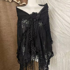 以下が含まれることがあります： マネキンに掛けられた黒いレースのショール。複雑な花柄、スカラップ状の縁取り、中央の布製フラワーモチーフが特徴です。裾には繊細なクロシェ編みのトリムと、垂れ下がるフリンジのディテールがあしらわれています。