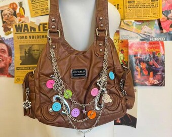 May include: Brown shoulder bag with decorative buttons in various colors and a silver chain. The bag has two pockets and the text "OUYALU" on the front. The bag is displayed on a white mannequin.