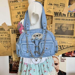 May include: A light blue handbag with silver chain straps and decorative charms. The bag features an eye-shaped embellishment and multiple pockets. The background includes vintage-style posters and a skirt with a cherry print.