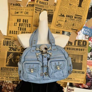 Könnte beinhalten: Hellblaue Handtasche mit Totenkopf- und gekreuzten Knochen-Verzierung. Die Tasche hat mehrere Taschen und dekorative Anhänger. Der Hintergrund zeigt Vintage-Zeitungsartikel mit Text wie "Ministry Decree" und "Harry Potter."