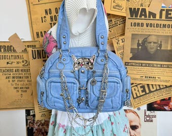 May include: A light blue handbag with a cat face patch, silver chains, and multiple pockets. The bag is displayed on a mannequin with a floral skirt and surrounded by vintage newspaper clippings and posters.