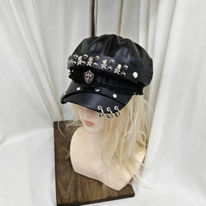 May include: A black faux leather newsboy cap featuring a decorative band adorned with silver-toned skull studs and round rivets. The front brim includes three dangling silver metal rings and a central shield-shaped emblem with a sword motif.