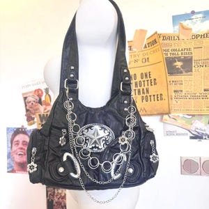 May include: Black faux leather handbag with silver-tone hardware. The bag features a star-shaped buckle, chain accents, and flower charms. The bag has two handles and a shoulder strap. The bag is displayed on a mannequin.
