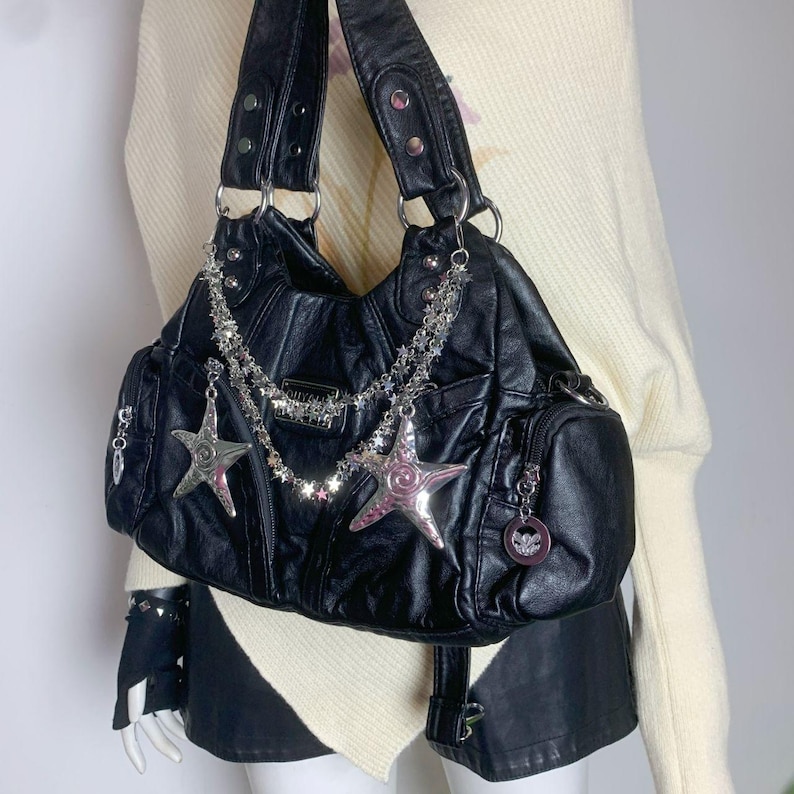May include: Black handbag with silver star and chain accents. The bag features multiple pockets and a textured finish. The bag has two handles and a silver chain with star charms. The bag is a shoulder bag.