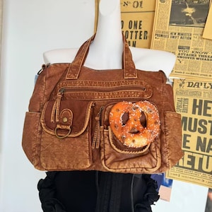 May include: A brown faux leather handbag with multiple pockets and zippers. A pretzel is placed in one of the front pockets. The bag has two handles and is displayed on a mannequin.
