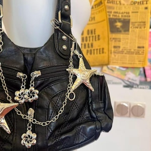 May include: Black leather shoulder bag with silver star and flower charms. The bag has a quilted pattern and multiple zippers. A silver chain and safety pin are attached. The background includes a yellow newspaper clipping.