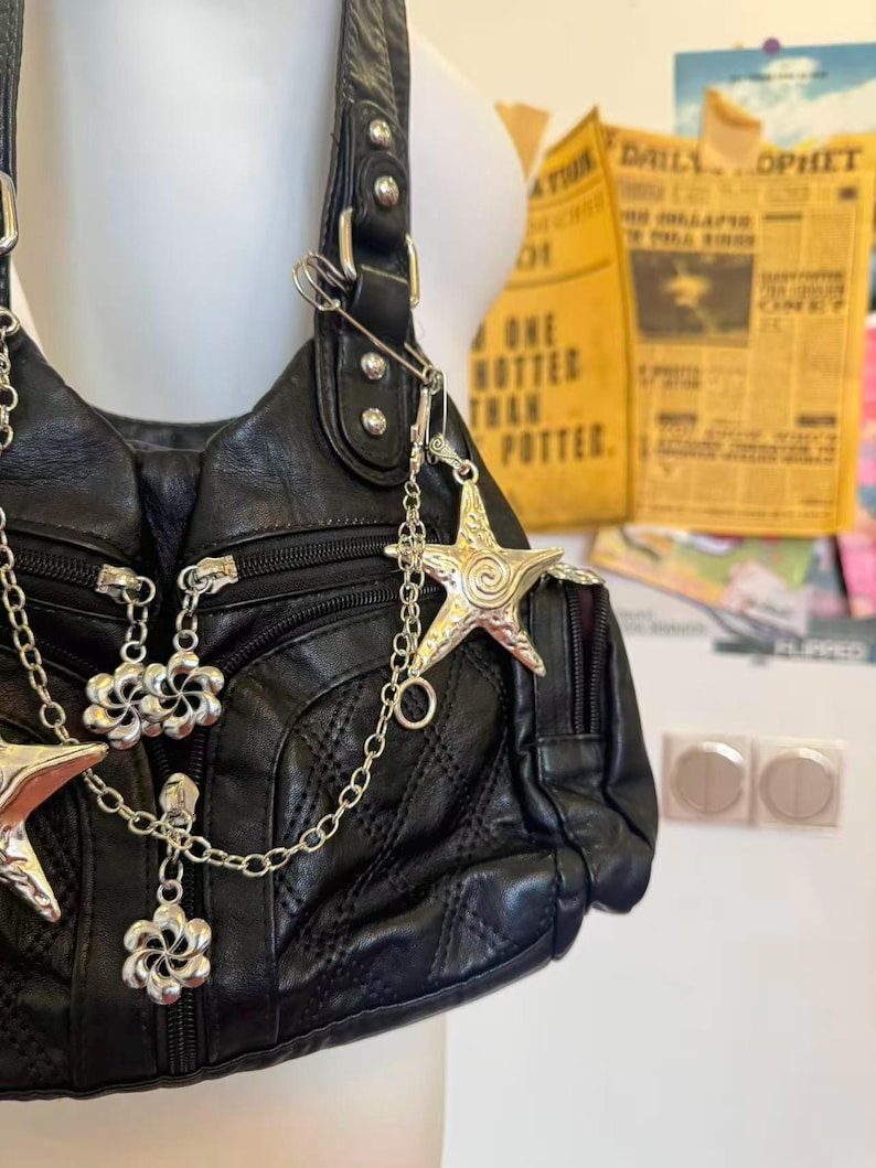 May include: Black leather shoulder bag with silver star and flower charms. The bag has a quilted pattern and multiple zippers. A silver chain and safety pin are attached. The background includes a yellow newspaper clipping.