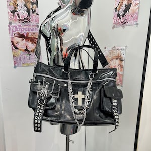 May include: A black bag featuring a silver cross emblem, a skeleton hand charm, and multiple silver chains. The bag includes two side pockets with grommet-detailed straps, a top zipper closure, and a wide shoulder strap with metal eyelets.