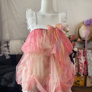 May include: A pink and white dress displayed on a mannequin. The dress features a white square neckline with ruffled trim, a pink and white layered skirt, and a bow. The dress is designed with a layered, tulle-like fabric.