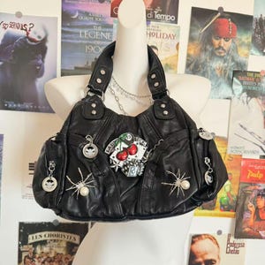 May include: Black leather handbag with silver detailing, adorned with a cherry and dice patch, spider charms, and a chain strap. The bag has multiple pockets and a top handle. Movie posters are in the background.
