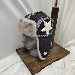 May include: A dark gray trapper hat featuring a beige faux fur lining and ear flaps. The hat is accented with a white fabric star patch, frayed edges, and a small round wooden button detail on the side, displayed on a gray mannequin head.