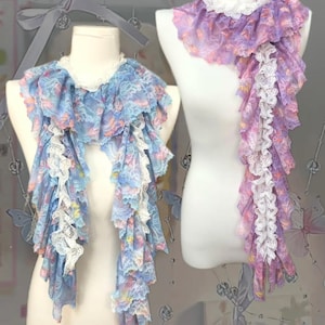 May include: Two decorative scarves on mannequins. One is pale blue with floral patterns and white lace trim. The other is purple with pink accents and white lace trim. Both have a layered, cascading design.