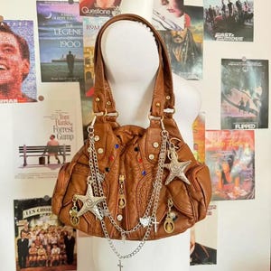 May include: A brown leather handbag with a ruched design, adorned with silver chains, star charms, and decorative studs. The bag has two handles and is displayed against a backdrop of movie posters.
