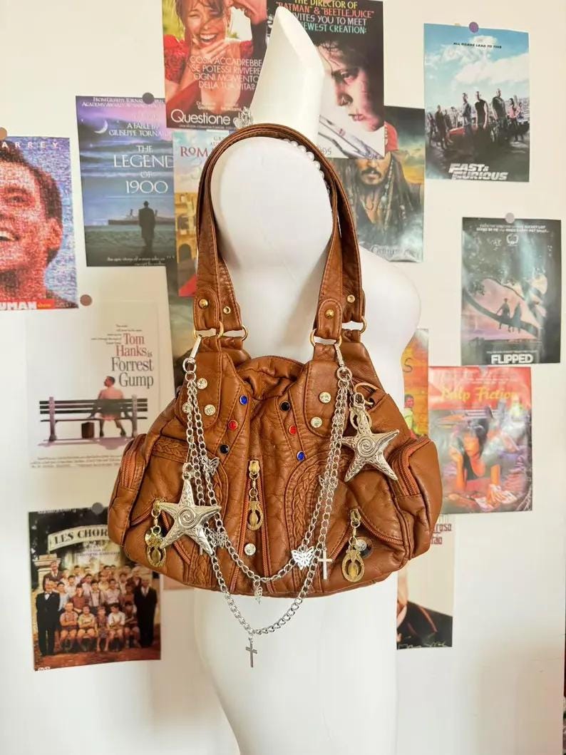 May include: A brown leather handbag with a ruched design, adorned with silver chains, star charms, and decorative studs. The bag has two handles and is displayed against a backdrop of movie posters.
