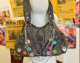 May include: Grey faux leather handbag with silver-tone hardware, decorative chains, and colourful buttons. The bag has multiple pockets and a top handle. Charms and star accents add detail to the design.