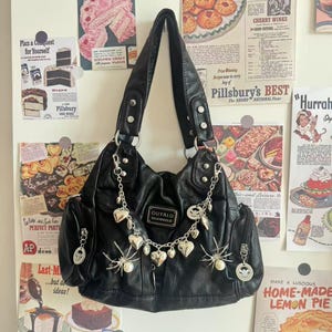 Könnte beinhalten: Schwarze Handtasche aus Kunstleder mit silberfarbenen Details. Die Tasche hat eine Kette mit Herz- und Spinnenanhängern und mehrere Taschen. Der Hintergrund zeigt Vintage-Lebensmittelwerbung.