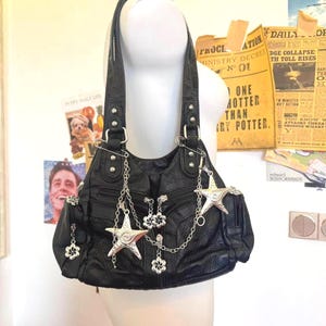 May include: Black leather shoulder bag with silver star and flower charms. The bag has a textured surface and multiple pockets. The bag is hanging from a mannequin.