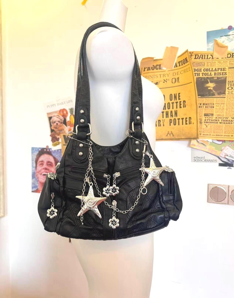May include: Black leather shoulder bag with silver star and flower charms. The bag has a textured surface and multiple pockets. The bag is hanging from a mannequin.