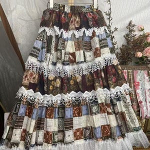 Peut inclure: Longue jupe patchwork avec une variété de motifs et de couleurs, dont des motifs floraux et géométriques. La jupe est ornée d'une bordure en dentelle blanche et d'un ourlet à volants. La jupe est suspendue à un cintre en bois.