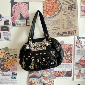 May include: Black leather handbag with a cat face design, embellished with pearls and rhinestones. The bag has gold-tone hardware and multiple pockets. Vintage-style advertisements are in the background, including a Pillsbury's Best ad.