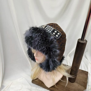 May include: A brown and black patterned hat featuring a thick, dark grey faux fur trim around the brim. The hat includes a black band across the top adorned with multiple clear, circular rhinestones arranged in a grid pattern for decoration.