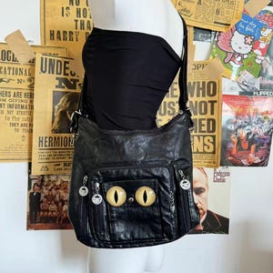 May include: Black leather shoulder bag with cat eye design. The bag features a large front pocket with two golden cat eyes and two smaller side pockets. The bag is displayed on a mannequin against a backdrop of vintage posters.