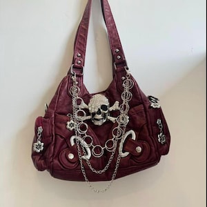 May include: A burgundy shoulder bag with a skull and crossbones design. The bag features a rhinestone-studded skull, silver chain accents, and flower charms. The bag has multiple pockets and a top handle.