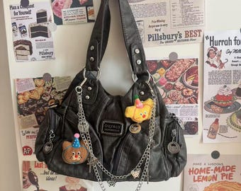 May include: A dark gray leather handbag with multiple pockets and silver chain accents. The bag features a bear and croissant pin, and a metal tag that reads "OUYALU". Vintage-style advertisements are in the background.