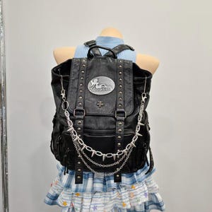Può includere: Uno zaino in similpelle nera con borchie metalliche color argento, una catena decorativa con punte e una placca metallica ovale sulla patta anteriore con la scritta "GOOD GIRL" e l'illustrazione di una figura distesa.