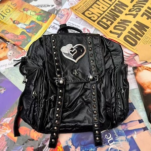 Puede incluir: Mochila negra con tachuelas plateadas, un emblema en forma de corazón y cierres con forma de araña. La bolsa tiene múltiples bolsillos y correas. El fondo incluye varios materiales impresos.