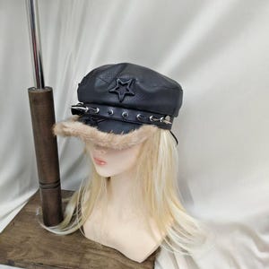 May include: A black faux leather military-style cap featuring a textured finish, a prominent star emblem on the front, and a black strap adorned with silver-toned metal spikes. The brim is trimmed with a light brown faux fur accent.