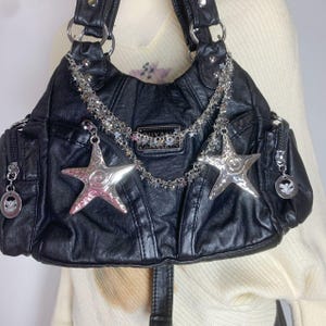 May include: Black faux leather handbag with silver star charms and chain accents. The bag features multiple pockets, silver hardware, and a decorative label. The bag has a shoulder strap and a zipper closure.