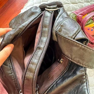 May include: A dark brown leather crossbody bag with multiple zipped compartments. The bag has a shoulder strap and silver-toned hardware. The interior lining is a lighter brown colour. The bag is open, revealing the interior.