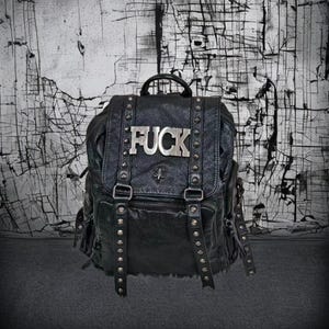 Puede incluir: Una mochila de cuero negro con superficie texturizada, tachuelas metálicas plateadas en las correas verticales y una placa de metal plateado en la solapa frontal con la palabra FUCK en letras mayúsculas. Incluye un asa superior.