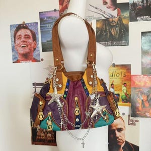 May include: A multi-colored handbag with brown handles and silver star and cross charms. The bag features purple, yellow, and teal panels with gold zippers and decorative stitching. The bag is displayed on a mannequin.