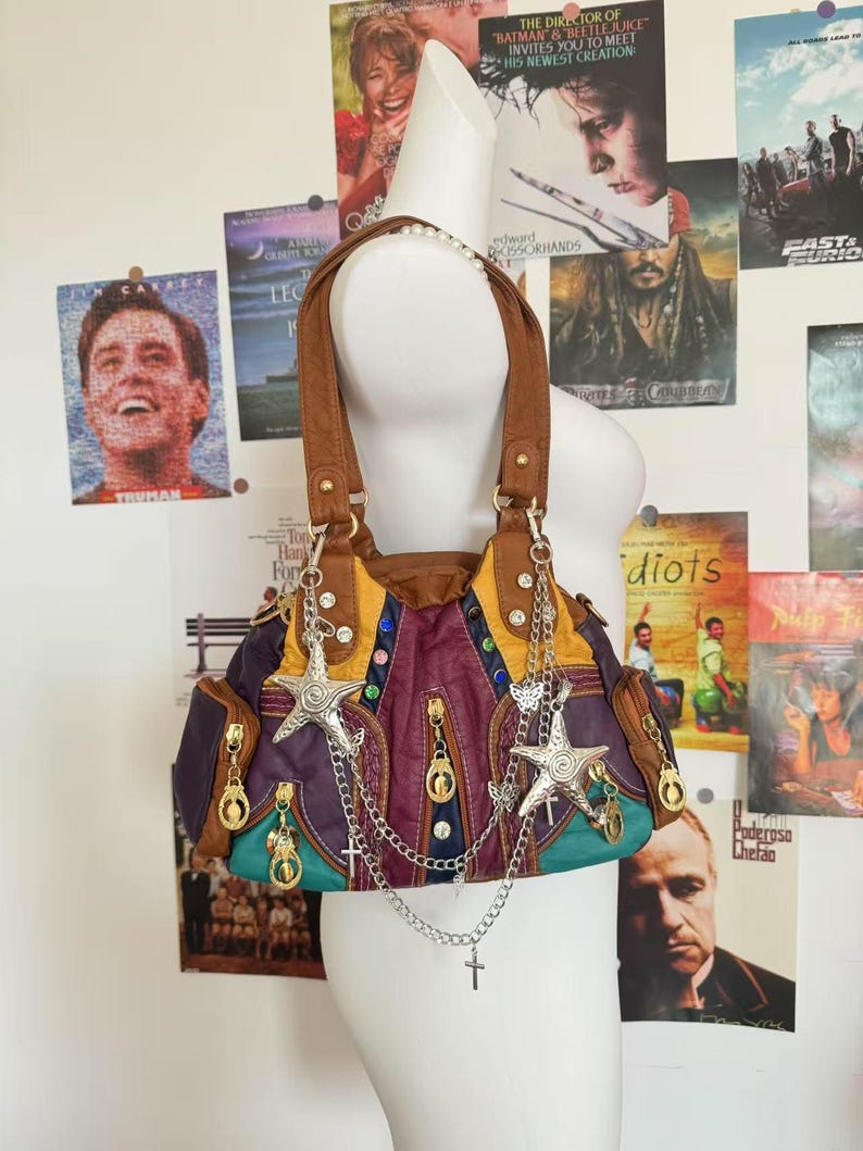 May include: A multi-colored handbag with brown handles and silver star and cross charms. The bag features purple, yellow, and teal panels with gold zippers and decorative stitching. The bag is displayed on a mannequin.