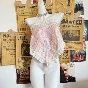 Puede incluir: Un busto blanco exhibe un top con tema de mariposa con un diseño floral rosa y blanco. El top presenta una aplicación de mariposa y ribete de encaje. Un collar plateado adorna el busto. El fondo incluye carteles y recortes de periódicos vintage.