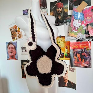 May include: A handmade, dark brown and cream-colored crochet flower bag. The bag is shaped like a large flower with a cream-colored center and trim. The strap is also cream-colored. The bag is displayed on a mannequin.