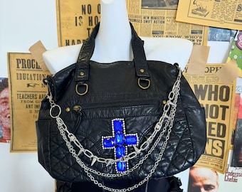 Könnte beinhalten: Schwarze Lederhandtasche mit Steppmuster, verziert mit einem blauen Juwelenkreuz und silbernen Kettenakzenten. Mit Tragegriffen, Schulterriemen und einer Fronttasche mit Reißverschluss. Die Tasche wird auf einer Schaufensterpuppe präsentiert.