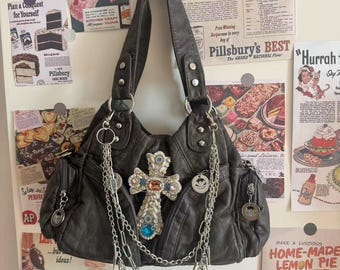 May include: Dark gray handbag with silver chain accents and a jeweled cross. The bag has multiple pockets and a decorative charm. Vintage-style advertisements are in the background.