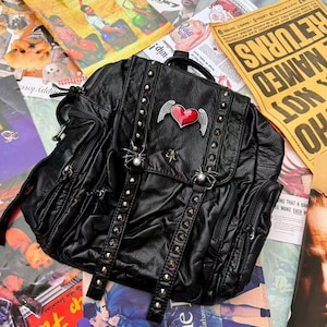 Puede incluir: Mochila negra de cuero sintético con tachuelas plateadas y un emblema de corazón con alas. La bolsa tiene múltiples bolsillos y correas, y está rodeada de coloridos carteles de películas y recortes de revistas.