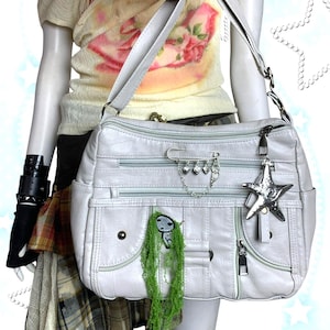 Pode incluir: Uma bolsa de ombro cinza claro com vários compartimentos com zíper e uma alça longa ajustável. A bolsa apresenta um pingente de estrela prateada e uma corrente decorativa com pingentes de coração. Um pequeno personagem verde e cinza está preso na frente.